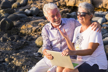 Wall Mural - Happy laughing and smiling caucasian cheerful adult mature couple having fun outdoor using technology laptop computer together under the sun in vacation sitting on the rocks