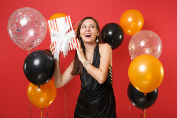 Funny girl in black dress celebrating, trying to guess what's in red box with gift, present on bright red background air balloons. Women's Day, Happy New Year, birthday mockup holiday party concept.