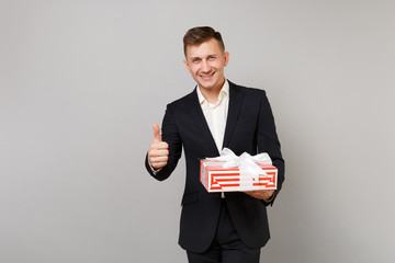 Joyful business man showing thumb up holding red striped present box with gift ribbon isolated on grey background. Achievement career wealth business. Valentine's Women's Day birthday holiday concept.