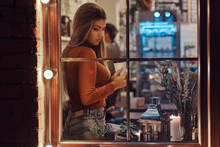 Beautiful Girl Wearing Sweater Holding A Coffee While Sitting On A Window Sill Inside The Cafe