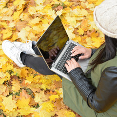 Wall Mural - Girl in hipster with laptop in autumn park. A woman in a cap using a laptop while sitting on fallen leaves. Freelancer in the hat uses remote communication technology. Remote work. View from above