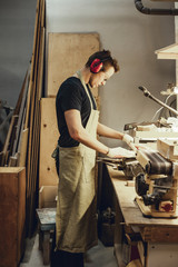 Wall Mural - Side view of young male in apron and earmuffs standing near workbench and using jigsaw while working in workshop