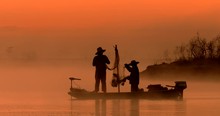 Two Silhouetted Traditional Asian Fishermen Using Nets To Fish At An Exotic River Location From A Flat-bottomed Boat At Sunrise Or Sunset