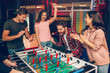 © estradaanton - Cheerful happy young team stand at soccer table in playing room and cheering. Bearded guy play with another human. Frieds surround him.