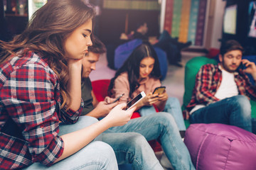 Bored young people sit in playing room and look at phoens in hands. One guy talk using cellphone. They busy.