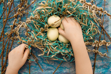 paper nest with three eggs with a kid's arms holding it and willow branches around them on a blue background