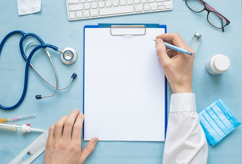 The top view of the doctor’s hands is writing down a prescription or medical report on a blank sheet of paper. On the table are stethoscope and other attributes. Copy space.  