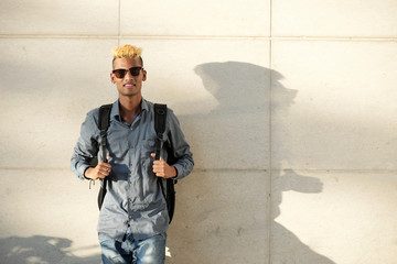 Portrait of cool young man in sunglasses standing at wall with big backpack