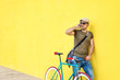 © Rafa Fernandez - Side view of a young adult man with a vintage bike and wearing casual clothes and sunglasses standing against a yellow wall while using a mobile phone in a sunny day