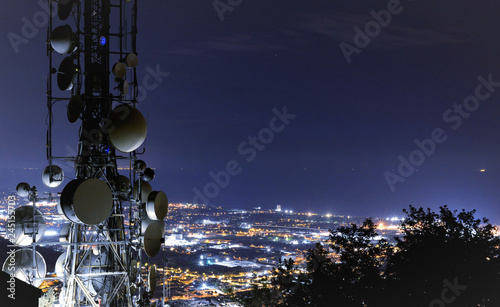 Fotografía Telecommunications tower, antenna and satellite dish and city at night as backgr