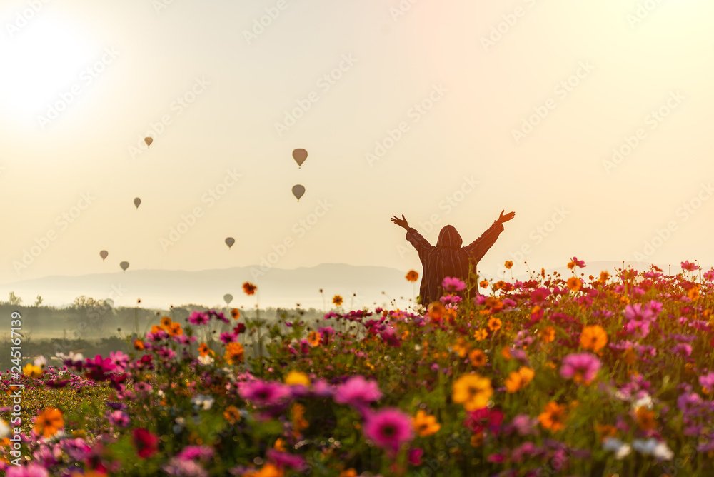 Lifestyle traveler women raise hand feeling good relax and happy freedom and see the fire balloon outdoors the nature tea and cosmos farm in the sunrise morning. Travel and summer Concept - obrazy, fototapety, plakaty 