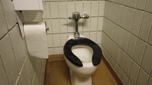 Public Washroom Toilet Stall With White Brick Walls, Silver Toilet Paper Holder And Plumbing And A Black Toilet Seat.