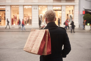 one young mans back, rear view, 20-29 years old, standing on a pedestrian street in city, looking, while holding two shopping bags on his back. Store front windows in distance.