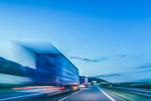 Background Photograph Of A Highway. Truck On A Motorway, Motion Blur, Light Trails. Evening Or Night Shot Of Trucks Doing Logistics And Transportation On A Highway.
