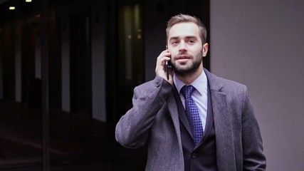 Poster - Smiling handsome business man in coat talking by smartphone while standing outdoors in the city