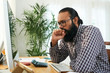 © DragonImages - Tense young programmer in casual shirt and eyeglasses sitting by desk in front of compter monitor while looking through data