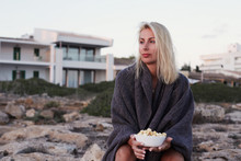 Authentic Portrait Of Young Dreaming Attractive Female With Blond Hair, Holding Popcorn, Going To Watch Sunset With Cozy Blanket On Shoulders, Thinking About Boyfriends, Sitting Outdoors On The Rocks.