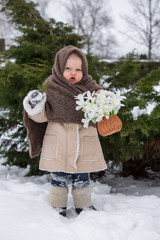 Winter among the trees a little girl of two years holding a snowdrop . The child is dressed in a sheepskin coat, shawl and felted boots. A very emotional child.