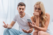 emotional man talking and looking at young wife using smartphone in bedroom, mistrust concept