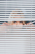 young woman looking at camera and peeking through blinds, mistrust concept