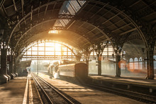 Old Railway Station With A Train And A Locomotive On The Platform Awaiting Departure. Evening Sunshine Rays In Smoke Arches.