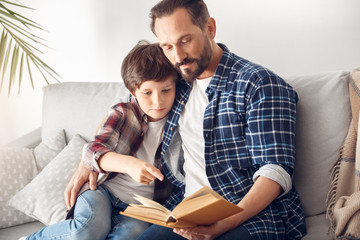 Wall Mural - Father and little son at home sitting on sofa hugging reading book together