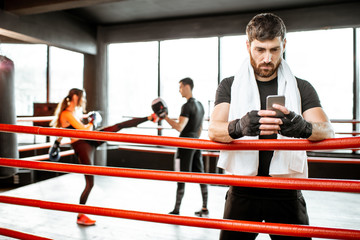 Wall Mural - Man resting with phone after the training on the boxing ring with people boxing on the background at the gym