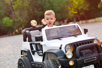 Cute 2 year old boy in a white T shirt is riding a white electric car in the park