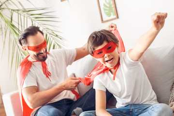 Wall Mural - Father and son in superheroe costumes at home sitting on sofa boy close-up hand up shouting motivated while dad holding his cloak happy