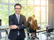 © kittipong - Successful asian young businessman with eyeglasses standing with arms crossed ,colleagues staff in background at office.