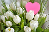 Fototapeta Tulipany - Bouquet of white beautiful tulips with tender petals and fresh green leaves with selective focus and blurred pink heart card for romantic valentines or mothers day. Spring bunch of flowers