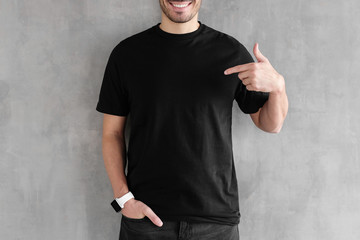 young man isolated on gray textured wall, smiling while pointing with index finger to black t-shirt,