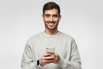 Wall Mural - Young man standing isolated on gray background, looking at camera with smile, holding smartphone with both hands