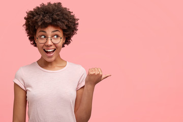 Horizontal view of happy mixed race teenager points with thumb at right side, concentrated aside, dressed in casual clothes, demonstrates something at blank empty space, expresses good emotions