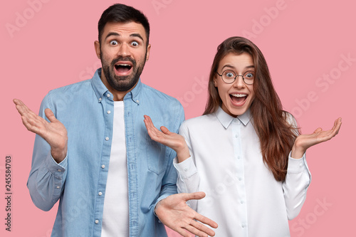 People, decision and attitude concept. Two hesitant business partners have glad facial expressions, shrug shoulders with uncertainty and bewilderment, isolated over pink background, express happiness