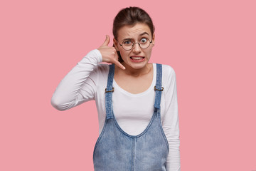 Wall Mural - Puzzled young woman clenches teeth, makes phone gesture, wears casual clothes, raises eyebrows in bewilderment, has round spectacles, stands against pink background, pretends calling someone.