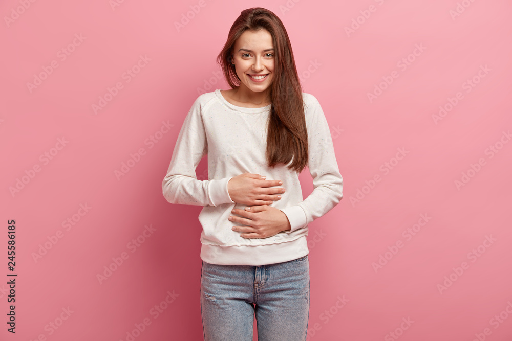 Satisfied smiling young woman keeps both hands on belly, being in good mood after eating delicious supper, demonstrates she is full, isolated over pink background. Pleasant feeling in stomach - obrazy, fototapety, plakaty 