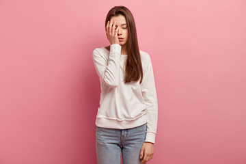 young woman has tired expression, covers part of face with palm, closes eyes, dressed in casual clot