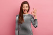 Photo of cheerful young woman blinks eye, makes okay gesture, demonstrates her agreement, feels happy, wears black and white striped jumper, isolated over pink background. Body language concept