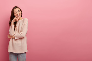 Wall Mural - Good looking young girl keeps hand under chin, focused aside with cheerful dreamy expression, feels pleased, dressed in casual sweater, models against pink studio wall, empty space for your promotion