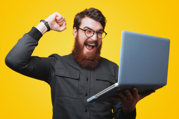 Wall Mural - Portrait of excited bearded man in glasses and gray shirt, is holding his laptop and gesturing victory