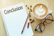 Conclusion word on notebook with glasses, pencil and coffee cup on wooden table. Business concept.