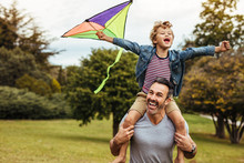 Smiling Boy On Fathers Shoulders Playing With Kite