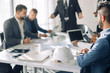© alfa27 - Close up shot of young business partners of construction compane sitting at table with white helmet on and listen as reporter man conducting a business presentation while standing in the board room.