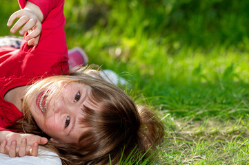 Cute pretty smiling child girl with gray eyes and long fair hair having fun outdoors laying on green grass on blurred sunny summer green field background. Beauty, dreams and games of childhood.