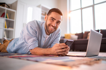 Wall Mural - Cheerful young man in casualwear lying on the floor of living-room in front of laptop and scrolling in smartphone