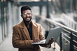 © F8  \ Suport Ukraine - Young indian bearded business man wear in coat and glasses standing outdoor with laptop in front of business building