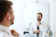 Smiling businessman putting on necktie while looking in the mirror and standing in the bathroom.