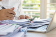 © NuPenDekDee - Teacher holding pen by hand for checking student homework assignments on desk in school office. Unfinished paperwork stacked in archive with color paper clips. Education and business concept.