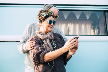 Cheerful old adult couple using technology smart phone internet to take selfie picture for social media accounts - cheerful people with old vintage blue van in background for travel and enjoy life